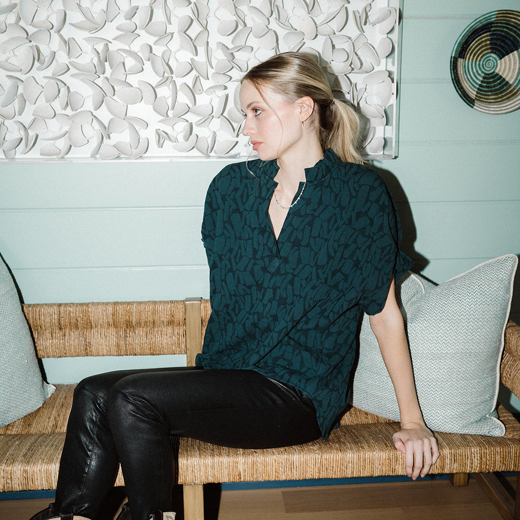 A woman sits on a woven bench in a styled room, wearing a dark teal patterned short-sleeve top, black leather pants, and cowboy boots. The background features a textured white wall panel and a light blue paneled wall.