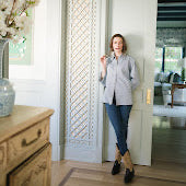 A woman stands in a doorway wearing a light gray button-up shirt, blue jeans, and black shoes, with patterned wallpaper and a wooden dresser visible in the room.